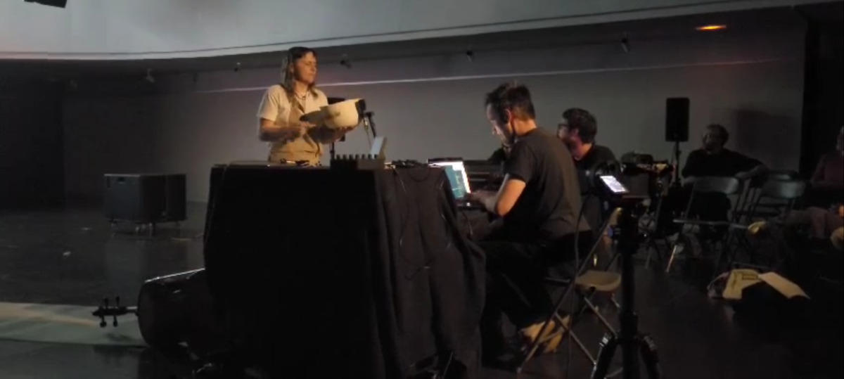 A man and a woman stand around a table in the middle of a concert space. The woman is hitting a ceramic bowl with a beater while the man types on a laptop.